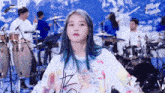a girl with blue hair is standing in front of a drum set and looking at the camera .