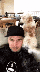 a man wearing a black hat is sitting on a couch with a small white dog sitting on his head