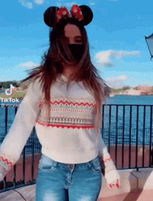 a woman wearing a mask and minnie mouse ears is dancing in front of a lake .