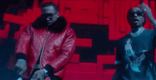 two men are standing next to each other on a stage . one of the men is wearing a red leather jacket .