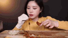 a woman in a yellow sweater and white gloves is eating chicken wings on a wooden table .
