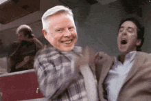 a man in a plaid shirt is smiling while another man in a suit is laughing