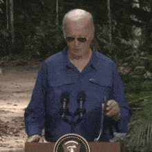 a man in a blue jacket is standing at a podium in front of microphones .