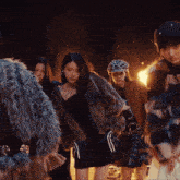 a group of women standing in front of a fire with one wearing a fur coat