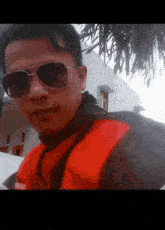 a man wearing sunglasses and a red jacket is standing in front of a house .