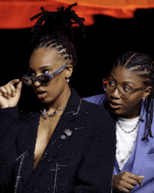 a woman wearing sunglasses stands next to another woman wearing a suit