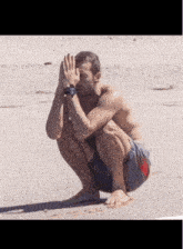 a shirtless man is squatting down on the beach