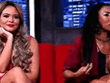 two women are sitting next to each other on a red couch and talking to each other .