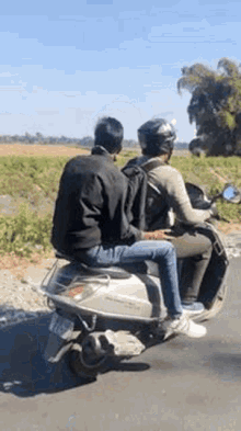 two men are riding a scooter down a road . one of the men is sitting on the back of the scooter .