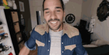 a man with a beard wearing a denim shirt is smiling in a room with a clock on the wall .