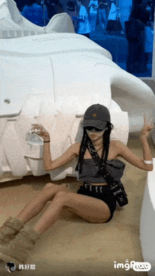 a woman in shorts and a hat is sitting on the ground in front of a large white object that says imgplay