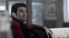 a man in a red sweater and black scarf is sitting on a couch and making a funny face .