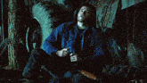 a man in a blue jacket sits on the ground holding a rifle