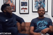 two men are sitting on a couch and one is wearing a kappa sweatshirt