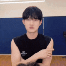 a young man in a black tank top is sitting in a room with his arms crossed and making a funny face .