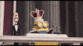 a minion with a crown on his head is standing in front of a microphone