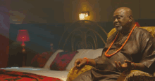a man wearing a red necklace is sitting in a chair in front of a bed
