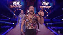 a man in a colorful shirt is pointing at the camera while standing on a stage in front of a rampage sign .