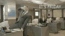 a man in a suit and tie is sitting at a desk in an office with the words `` it 's friday '' above him .