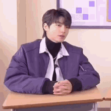 a young man in a purple jacket is sitting at a desk in a classroom with his hands folded .