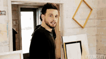 a man with a beard is standing in a doorway next to a stack of frames .