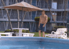 a shirtless man in blue shorts is standing next to a swimming pool under an umbrella