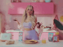 a woman in a purple top is standing in a kitchen with pink cabinets holding a spoon .