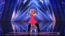 a little girl in a red dress is dancing with a man on a stage with a nbc logo in the background