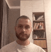 a man with a beard wearing a white shirt is standing in front of a bookshelf