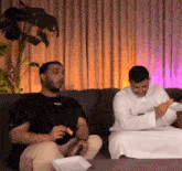 two men are sitting on a couch eating chips and one of them is wearing a black shirt that says stay free