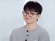 a young man wearing glasses and a gray shirt smiles for the camera with chinese writing behind him