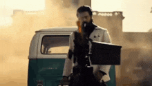 a man in a white suit is holding a gun in front of a green van .