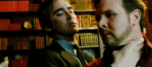 two men are standing next to each other in front of a bookshelf . one of the men is holding the other 's neck .