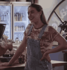 a woman wearing overalls is smiling in front of a fridge full of bottles