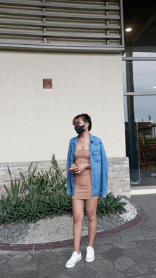 a woman wearing a face mask and a denim jacket stands in front of a building