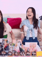 two women are sitting on a couch laughing in front of a table with a display of dolls on it