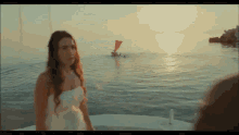 a woman in a white dress stands on a boat in the ocean with a red sailboat in the background