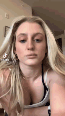 a woman with long blonde hair and blue eyes is wearing a white tank top and black bra .