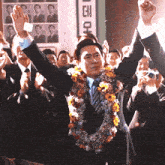 a man in a suit with a flower garland around his neck stands in front of a crowd with his arms in the air