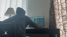 a man in a hoodie sits at a desk using a computer