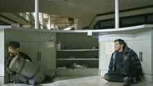 two men are sitting on the floor in an empty room with a sign on the wall that says ' emergency '