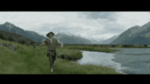 a man in a hat is walking across a grassy field next to a river