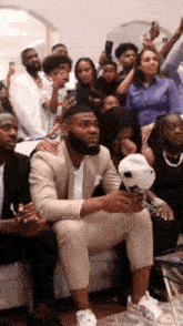 a man in a suit is sitting in front of a crowd of people holding a baseball cap .