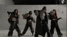 a group of women are dancing in front of a wall and one of them is wearing a fur coat