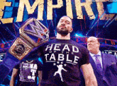 a man in a head of the table t-shirt holds a wrestling championship belt