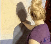 a woman wearing a purple shirt with a blue arrow on the back is touching her shadow