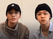 two young men wearing baseball caps and beanies are sitting next to each other and looking at the camera .