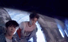 two young men are standing next to each other in front of an airplane . one of the men is wearing a punk shirt .