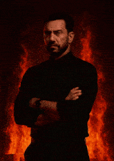 a man in a black shirt and tie stands in front of a fire