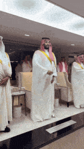 a man in a white robe with a yellow sash around his neck stands in a room with other men